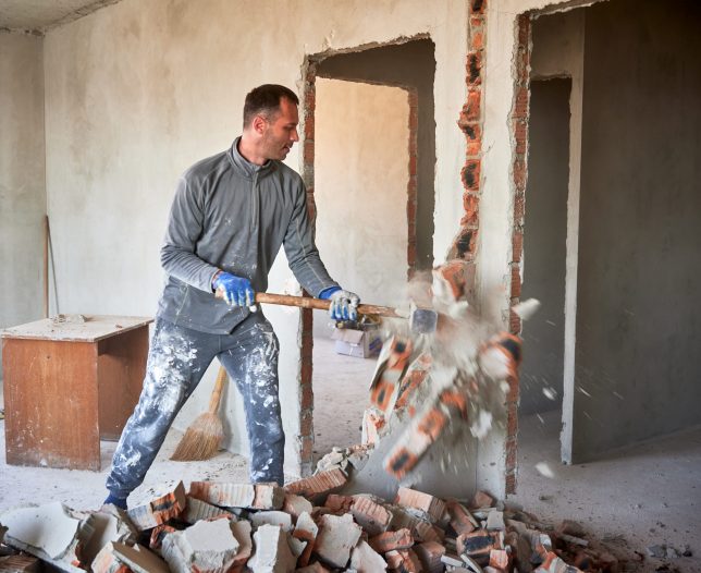 Repairman crashing brick wall with sledgehammer, pieces of brick flying away from strong blow. Destruction of partitions in flat during overhaul.