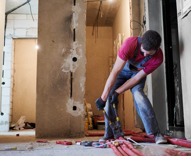 Full length of young man in work overalls connecting pipe with special tool. Male plumber in gloves using pipe tool while installing heating system in apartment. Concept of heating system installation