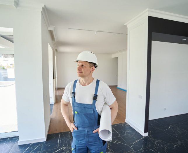 Adult caucasian male worker or architect with hands in pocket holding blueprint and looking away in new modern comfortable townhouse before repair