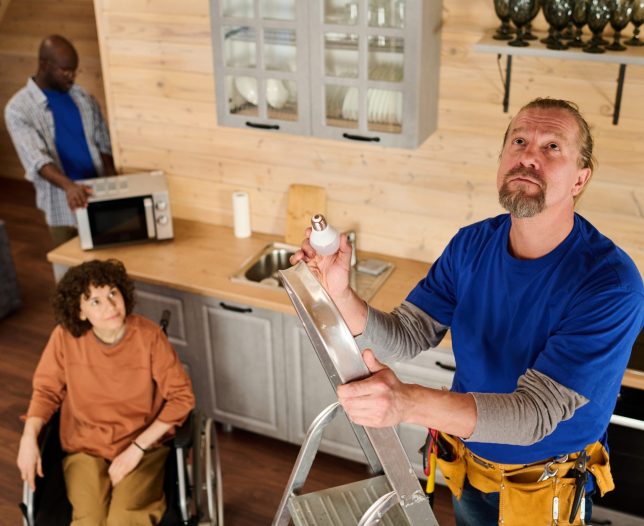 Mature bearded electrician with bulb standing on stepladder and looking upwards against woman in wheelchair and his colleague